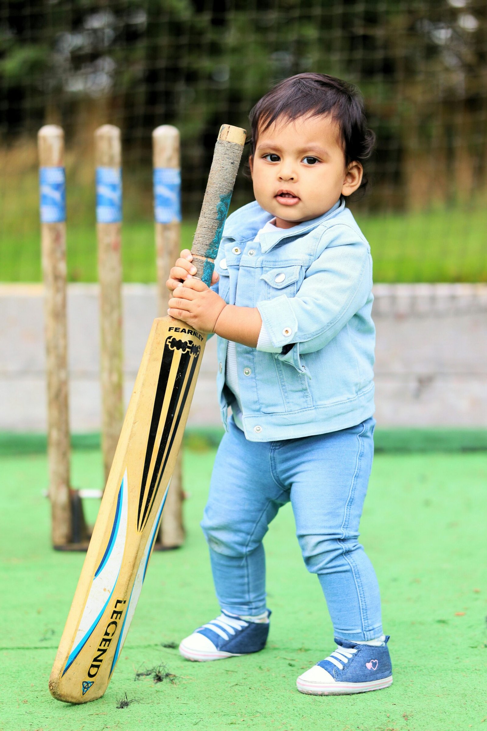Cute toddler in denim holding a cricket bat on a playing field.